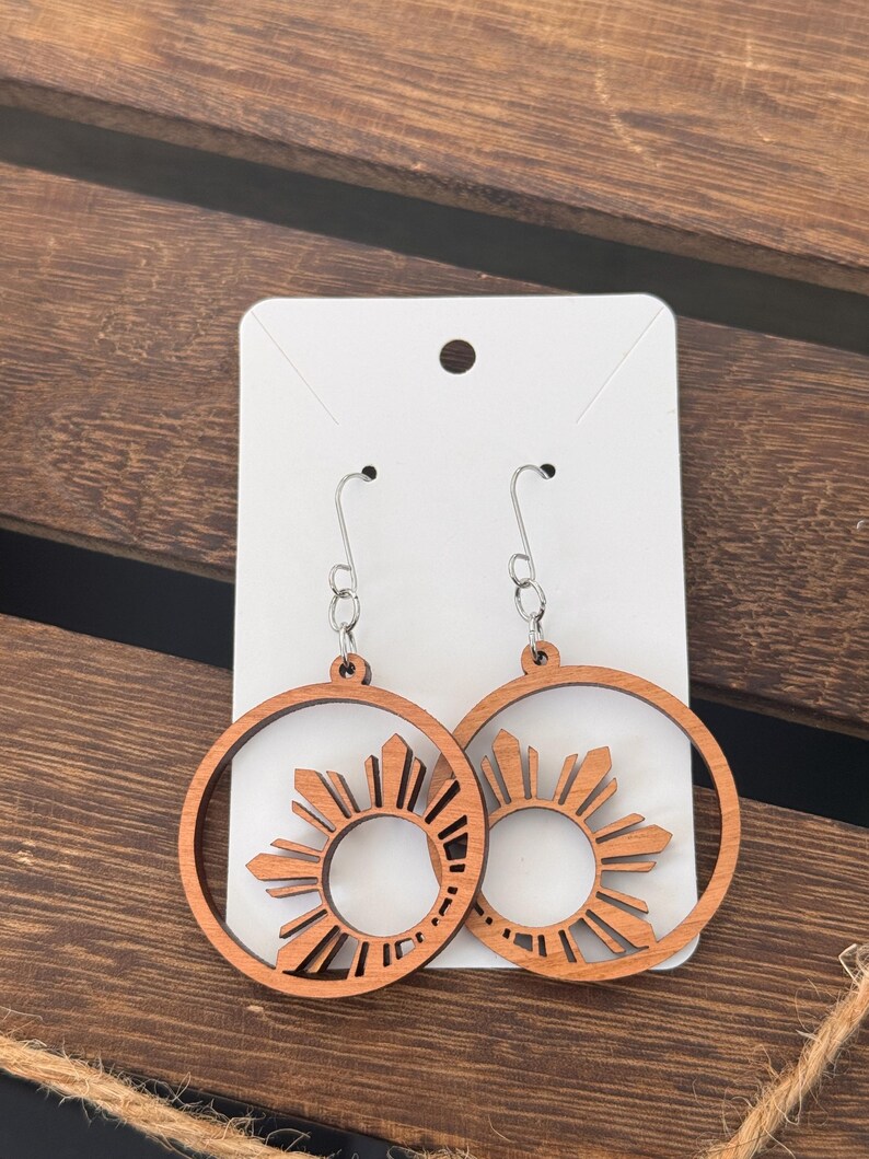 May include: A pair of wooden earrings with a sun design. The earrings are brown and have a silver hook.