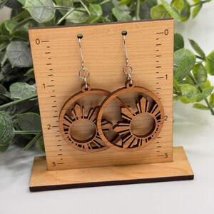 May include: A pair of wooden earrings with a sun design. The earrings are about 2 inches in diameter and are displayed on a wooden stand with a ruler.