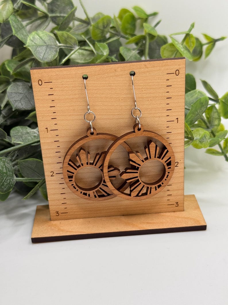May include: A pair of wooden earrings with a sun design. The earrings are about 2 inches in diameter and are displayed on a wooden stand with a ruler.