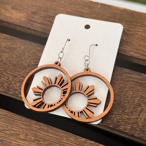 May include: A pair of wooden earrings with a sun design. The earrings are brown and have silver hooks.