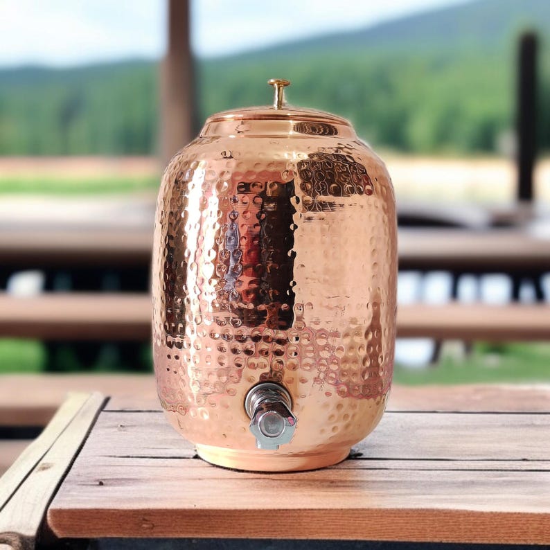 A hammered copper water dispenser with a spigot. The dispenser is a light copper color and has a round shape. The dispenser is sitting on a white surface.