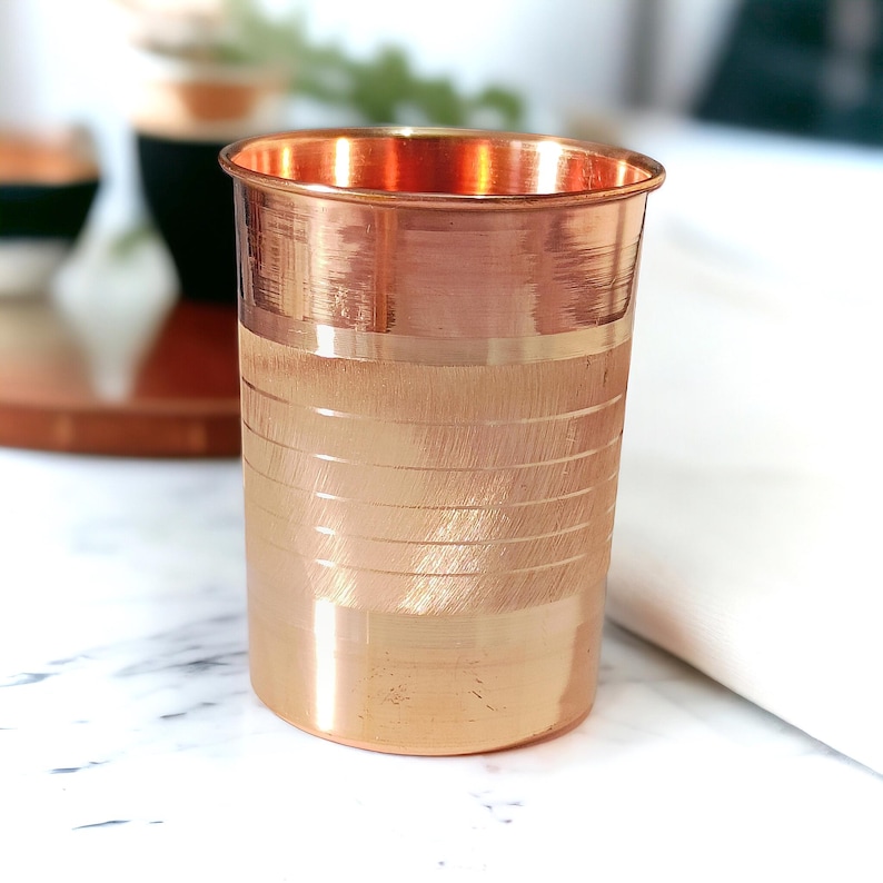 May include: A copper-colored drinking glass with a cylindrical shape. The glass has a polished rim and a textured, horizontal-lined design on the lower portion. The glass is made of copper and has a shiny, reflective surface.