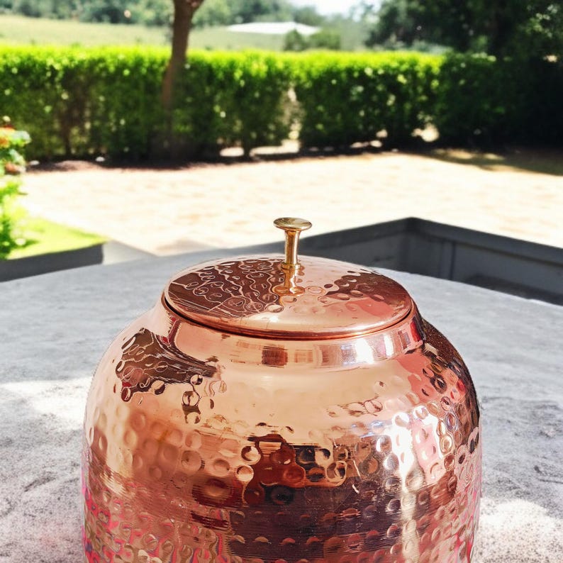 A hammered copper water dispenser with a spigot. The dispenser is a light copper color and has a round shape. The dispenser is sitting on a white surface.