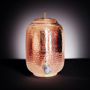 A hammered copper water dispenser with a spigot. The dispenser is a light copper color and has a round shape. The dispenser is sitting on a white surface.
