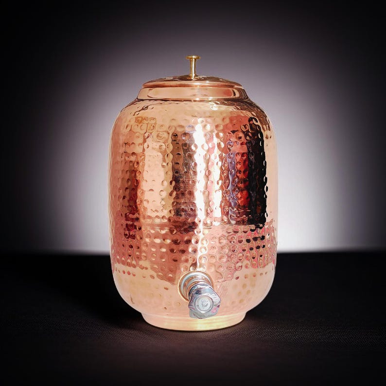 A hammered copper water dispenser with a spigot. The dispenser is a light copper color and has a round shape. The dispenser is sitting on a white surface.