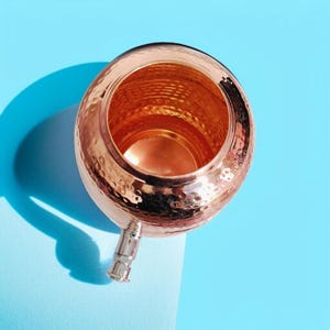 A copper water dispenser with a hammered finish. The dispenser has a spout and a lid. The dispenser is on a blue surface.