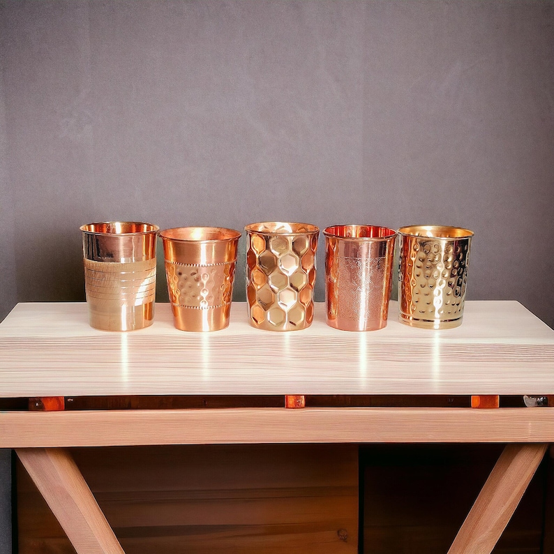 May include: Five copper-colored drinking glasses with varying textures are displayed on a light wood surface. The textures include horizontal lines, dimples, and a honeycomb pattern. The glasses are cylindrical with a slightly flared rim.