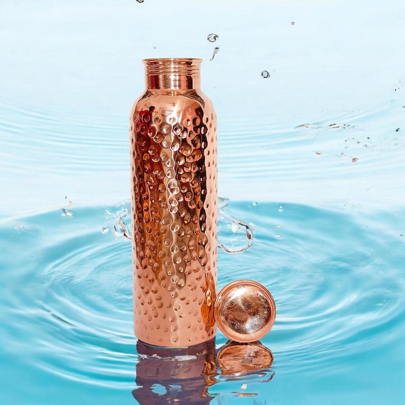 May include: A copper water bottle with a hammered finish. The bottle is sitting on a blue surface with water ripples.