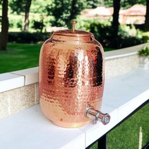 A hammered copper water dispenser with a spigot. The dispenser is a light copper color and has a round shape. The dispenser is sitting on a white surface.