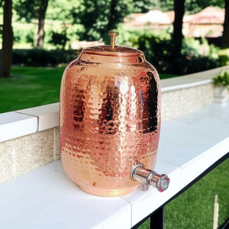 A hammered copper water dispenser with a spigot. The dispenser is a light copper color and has a round shape. The dispenser is sitting on a white surface.
