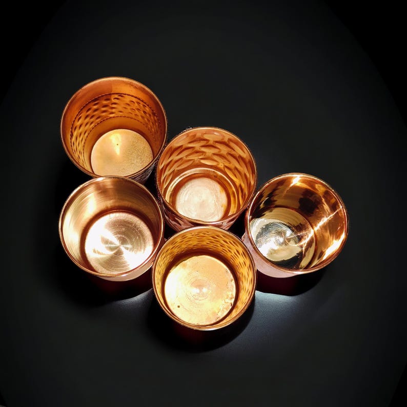 May include: Five copper-colored metal votive candle holders arranged in a circular pattern. The holders have a shiny, reflective surface, with some featuring a textured, patterned design. The background is a dark, solid color.