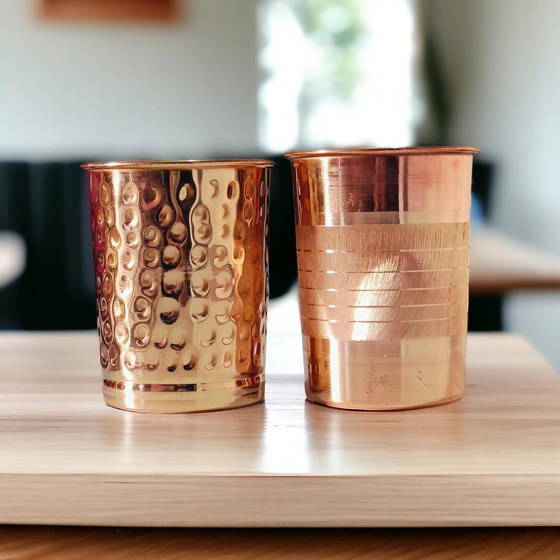 May include: Two copper drinking glasses on a wooden surface. One glass has a hammered texture, and the other has horizontal lines. The glasses are cylindrical and have a shiny, metallic finish. The background is blurred, with a glimpse of a window and a table.