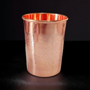 May include: A copper-colored drinking glass with an etched floral pattern. The glass has a smooth, polished exterior and a slightly flared rim. The interior of the glass has a bright, reflective finish.