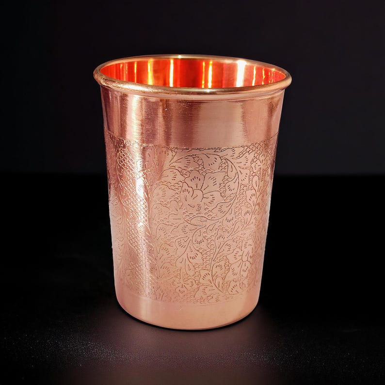 May include: A copper-colored drinking glass with an etched floral pattern. The glass has a smooth, polished exterior and a slightly flared rim. The interior of the glass has a bright, reflective finish.