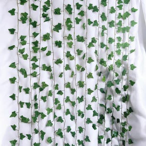 Puede incluir: Vides de hiedra artificiales verdes con pequeñas hojas detalladas dispuestas verticalmente sobre un fondo blanco. Las finas vides marrones crean una apariencia natural. Las hojas son de un verde vibrante, agregando un toque de naturaleza.