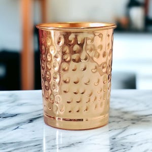 May include: A copper-colored tumbler with a hammered texture. The cylindrical cup has a wide mouth and a slightly flared rim. The surface is covered in small, evenly spaced dimples, giving it a unique, handcrafted appearance. The cup sits on a marble surface.
