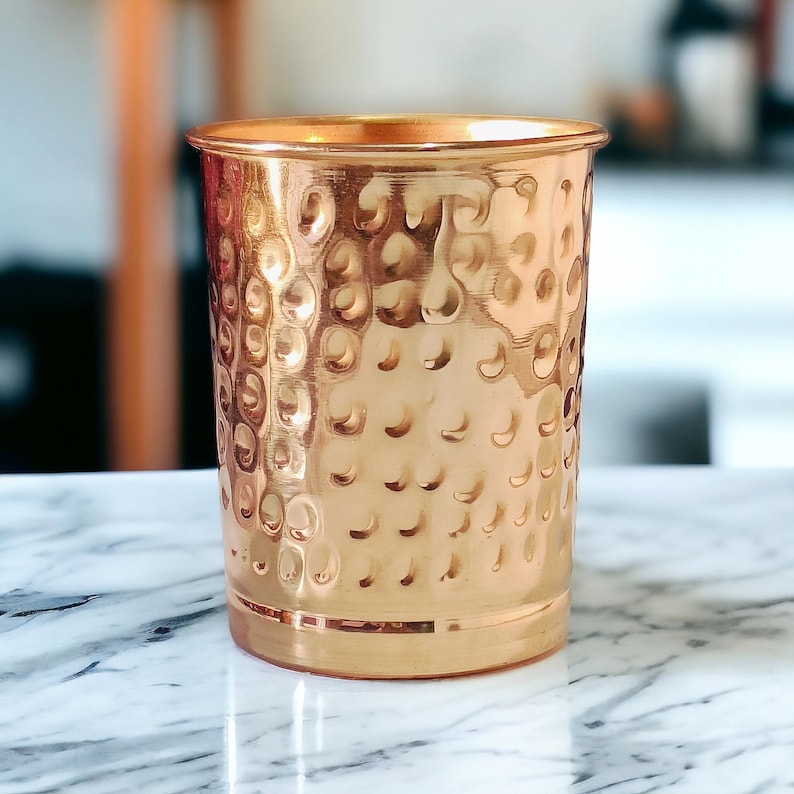 May include: A copper-colored tumbler with a hammered texture. The cylindrical cup has a wide mouth and a slightly flared rim. The surface is covered in small, evenly spaced dimples, giving it a unique, handcrafted appearance. The cup sits on a marble surface.