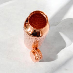May include: A copper water bottle with a hammered finish and a lid. The bottle is empty and the lid is off.