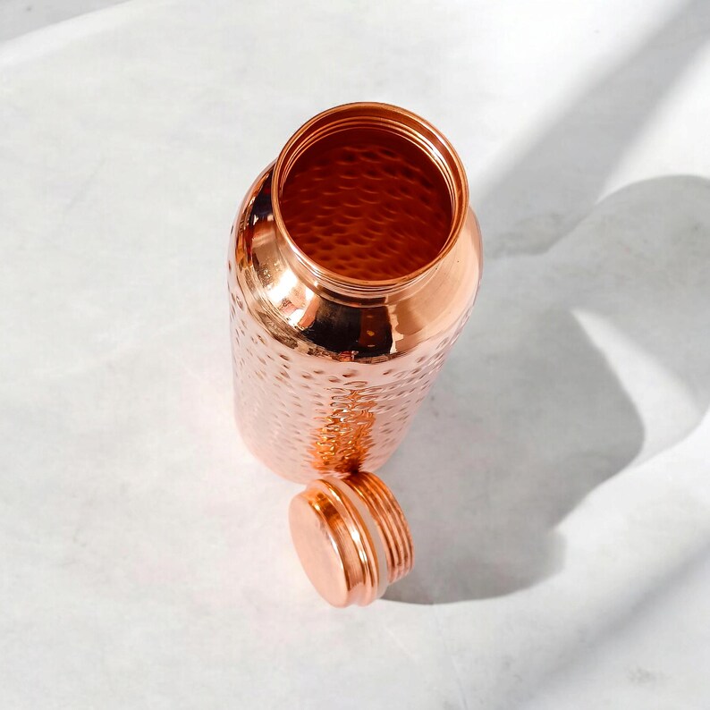 May include: A copper water bottle with a hammered finish and a lid. The bottle is empty and the lid is off.
