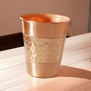 May include: A copper-colored metal tumbler with a hammered texture on the lower half. The top and bottom have a smooth finish, and the rim is slightly flared. The tumbler sits on a light-colored wooden surface.