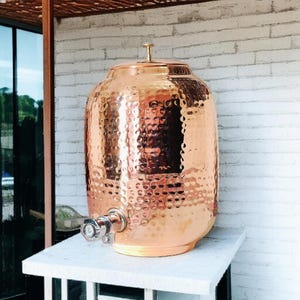 May include: A hammered copper water dispenser with a spigot. The dispenser is on a white marble surface.