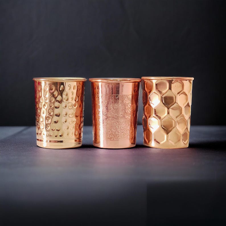 May include: Three copper drinking glasses with different textured surfaces. One has a hammered dimpled pattern, another has an etched design, and the third has a honeycomb pattern. The glasses are cylindrical and have a shiny finish.