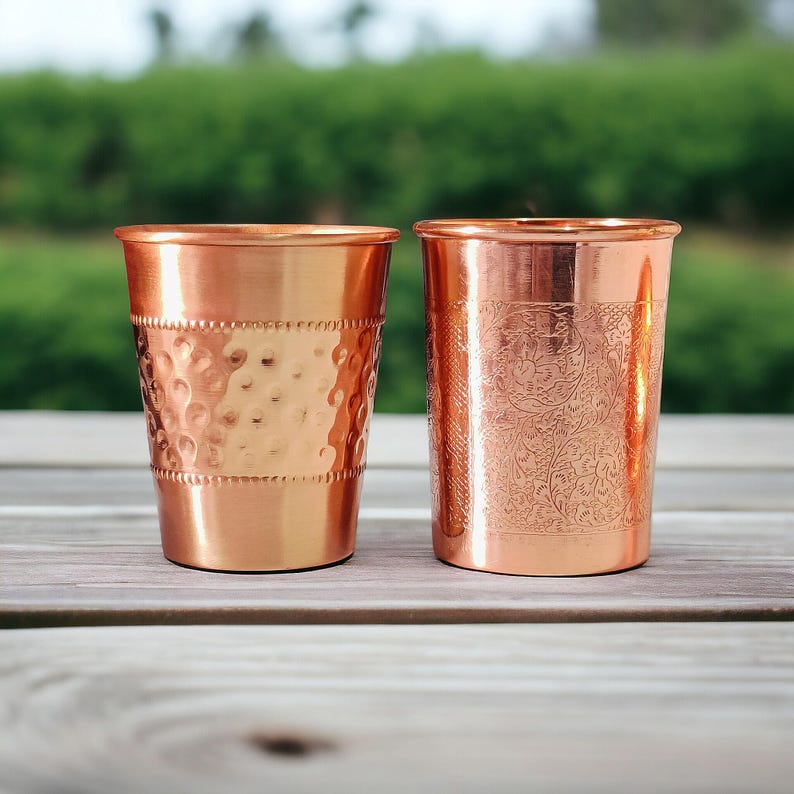 May include: Two copper drinking glasses. One has a hammered texture, and the other has an etched floral design. Both glasses are cylindrical with a polished finish. They sit on a wooden surface with a blurred green background.