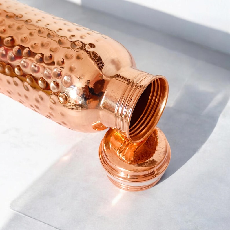May include: A copper water bottle with a hammered finish and a matching lid. The bottle is open and the lid is sitting next to it.