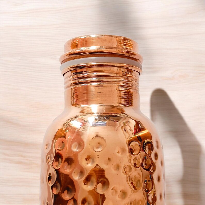 May include: A hammered copper water bottle with a copper lid and a white rubber seal. The bottle has a shiny, metallic finish.