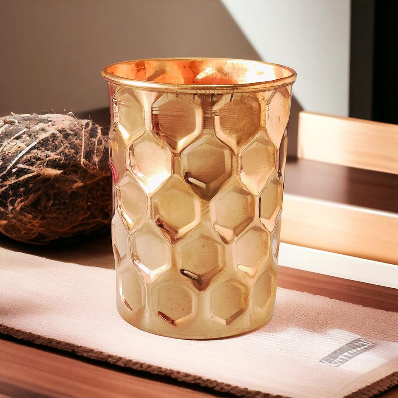 May include: A gold-colored glass with a honeycomb pattern. The glass has a cylindrical shape and a slightly flared rim. The surface reflects light, creating a warm, inviting glow. The glass is sitting on a white cloth.