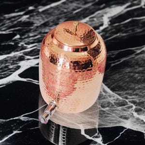 A hammered copper water dispenser with a spigot. The dispenser is a light copper color and has a round shape. The dispenser is sitting on a white surface.