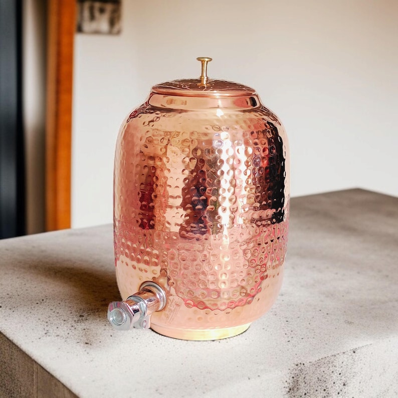 A hammered copper water dispenser with a spigot. The dispenser is a light copper color and has a round shape. The dispenser is sitting on a white surface.
