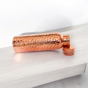 May include: A copper water bottle with a hammered finish and a lid. The bottle is lying on a light wood surface.