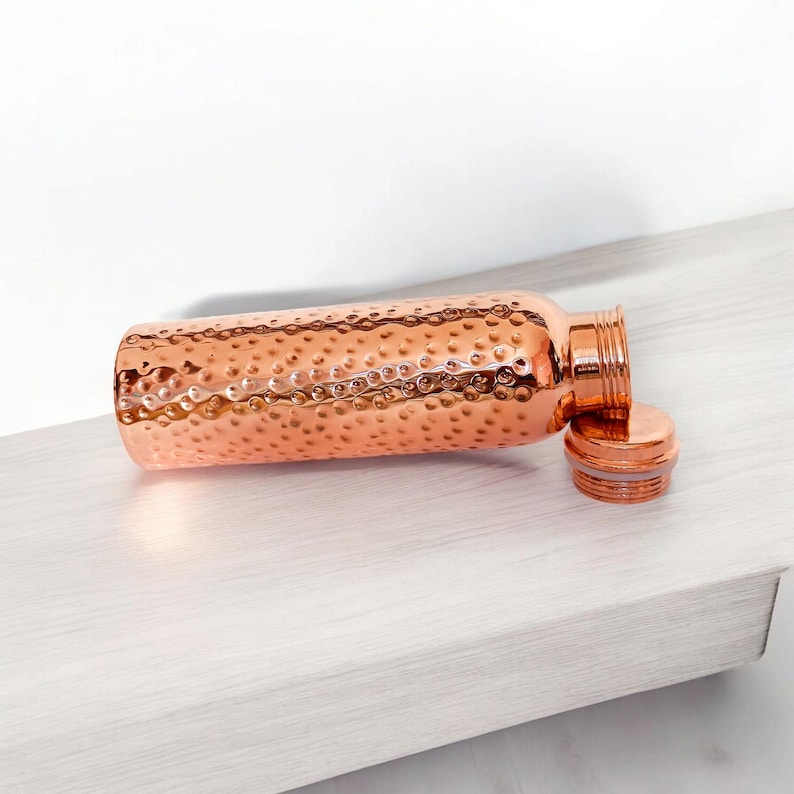 May include: A copper water bottle with a hammered finish and a lid. The bottle is lying on a light wood surface.