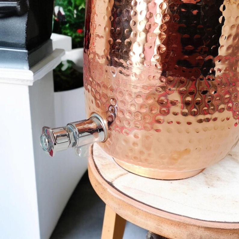 A hammered copper water dispenser with a spigot. The dispenser is a light copper color and has a round shape. The dispenser is sitting on a white surface.