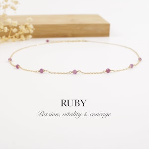 May include: A delicate gold necklace featuring small, round, ruby gemstones evenly spaced along the chain. The word "RUBY" is displayed below the necklace, along with the words "Passion, vitality & courage". The necklace is set against a white background.