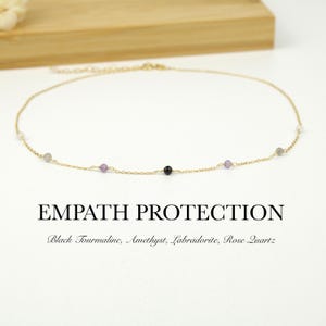 May include: A fine gold chain necklace adorned with small, colourful gemstone beads. The necklace includes black tourmaline, amethyst, labradorite, and rose quartz. The text "EMPATH PROTECTION" is displayed below the necklace.