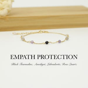 May include: A gold bracelet with a chain design, adorned with various gemstones in shades of purple, black, and pink. The bracelet is presented on a white surface with the text "EMPATH PROTECTION" and the names of the gemstones.