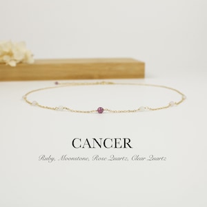 Puede incluir: Delicado collar de oro con un pequeño rubí y varias piedras blancas. La palabra "CANCER" se muestra debajo, junto con los nombres de las piedras: Ruby, Moonstone, Rose Quartz, Clear Quartz.