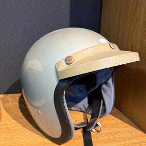 Könnte beinhalten: Silberfarbener Motorradhelm mit beigefarbenem Visier. Der Helm hat einen schwarzen Rand um die Öffnung und einen schwarzen Kinnriemen. Das Visier ist mit zwei kleinen, runden, rostfarbenen Nieten befestigt.