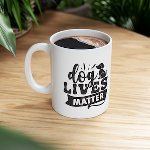 Op de afbeelding: Een witte keramische koffiemok met een zwarte handgreep. De mok heeft een zwarte afbeelding met de tekst "dog lives matter" en een klein zwart silhouet van een hond die op het woord "dog" zit.