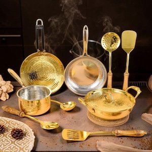 May include: A collection of gold-toned hammered metal cookware and utensils, including two frying pans, a small saucepan, a lidded pot, a slotted spoon, and two spatulas with wooden handles, arranged together with wisps of steam rising above.