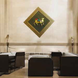 May include: A gold-colored, diamond-shaped wall art piece featuring a golden bull with colorful accents. The bull is centered on a dark green background within a gold frame. The artwork is displayed on a cream-colored wall.