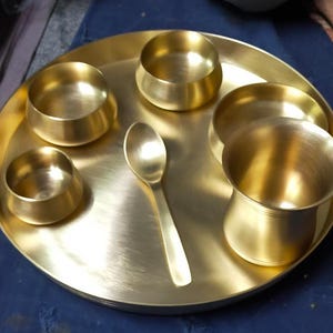 May include: A golden brass thali set, comprising a large round plate, five small bowls, and a spoon. The thali is a traditional Indian serving platter. The set is made of polished brass, ideal for presenting a variety of dishes.