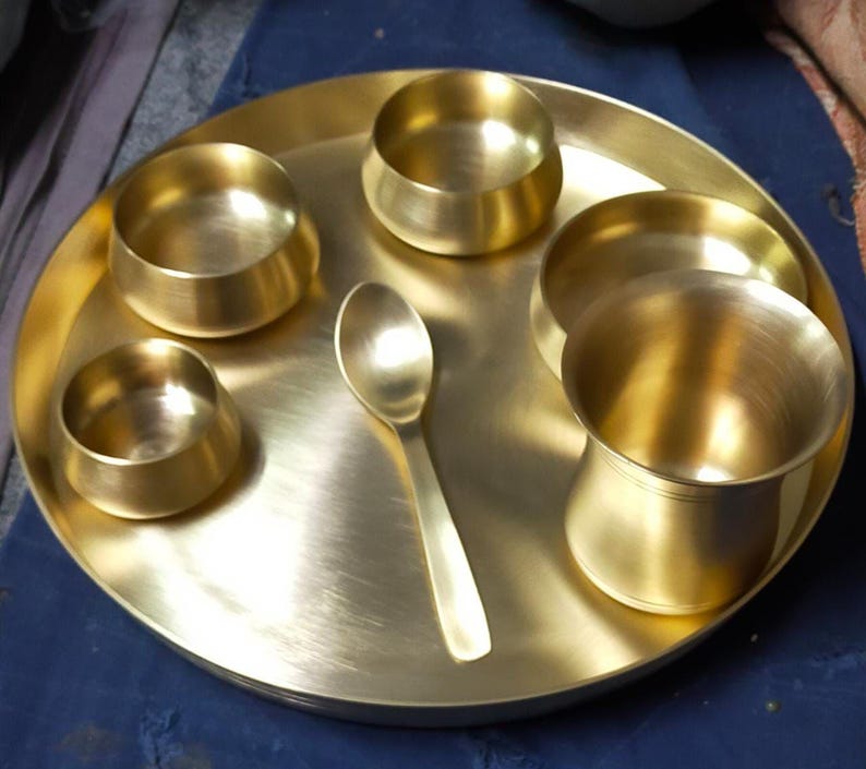 May include: A golden brass thali set, comprising a large round plate, five small bowls, and a spoon. The thali is a traditional Indian serving platter. The set is made of polished brass, ideal for presenting a variety of dishes.