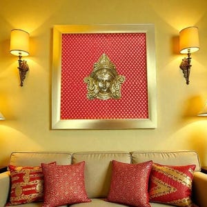 Puede incluir: Una escultura dorada y ornamentada de una cara está montada sobre una tela roja estampada dentro de un marco dorado. La obra de arte cuelga sobre un sofá beige con cojines rojos y dorados. Dos apliques de pared con pantallas flanquean la obra.