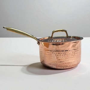 Peut inclure: Casserole en cuivre avec une finition martelée et un couvercle. La casserole a une longue poignée dorée et une petite poignée sur le couvercle. Le pot en cuivre est conçu pour la cuisine et a un aspect classique et élégant.