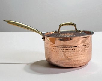 May include: Copper saucepan with a hammered finish and a lid. The pan has a long, gold-coloured handle and a small handle on the lid. The copper pot is designed for cooking and has a classic, elegant look.