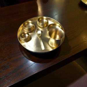 May include: A brass thali set, featuring a round tray, small bowls, a cup, and a spoon. The set is made of shiny, golden-coloured metal and is arranged on a dark wooden surface. The thali is a traditional Indian serving plate.