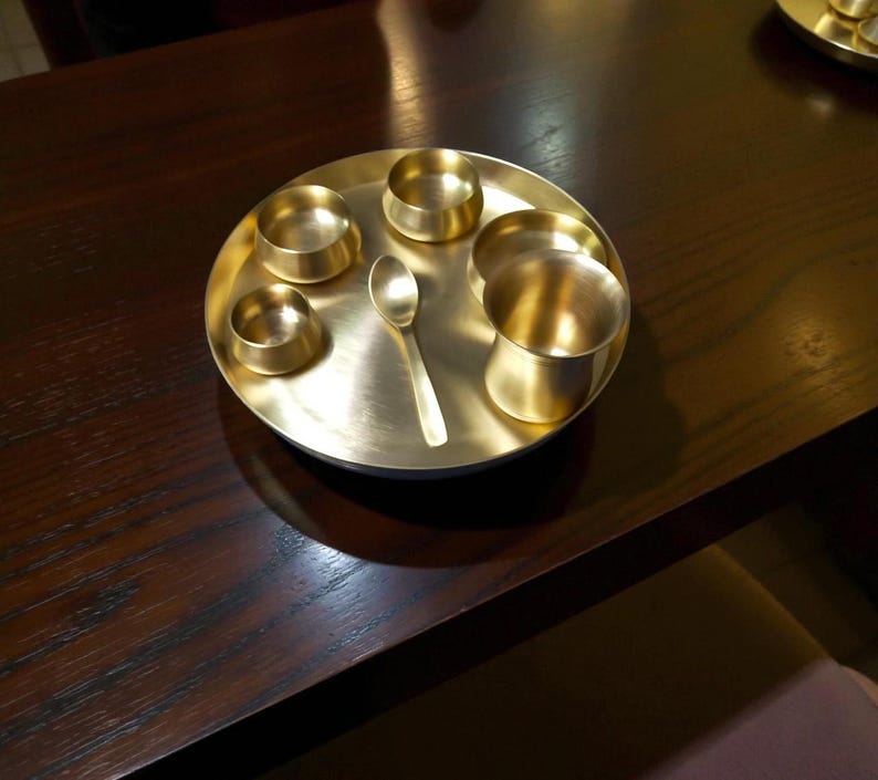 May include: A brass thali set, featuring a round tray, small bowls, a cup, and a spoon. The set is made of shiny, golden-coloured metal and is arranged on a dark wooden surface. The thali is a traditional Indian serving plate.